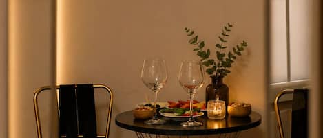 A hotel room with a bed, a bedside table with a smartphone, a small round table with a plant, two wine glasses, and a candle.