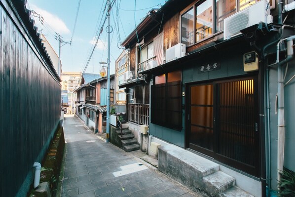 A narrow alleyway with traditional Japanese buildings, wooden doors, and a small staircase leading to a second-floor entrance.