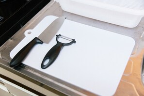 A kitchen counter with a white cutting board, a large knife, and a black peeler.