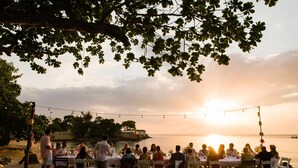 An outdoor dining area with tables and chairs arranged under a tree, overlooking a body of water during sunset. - Beachcliff Villa Negril (Negril)