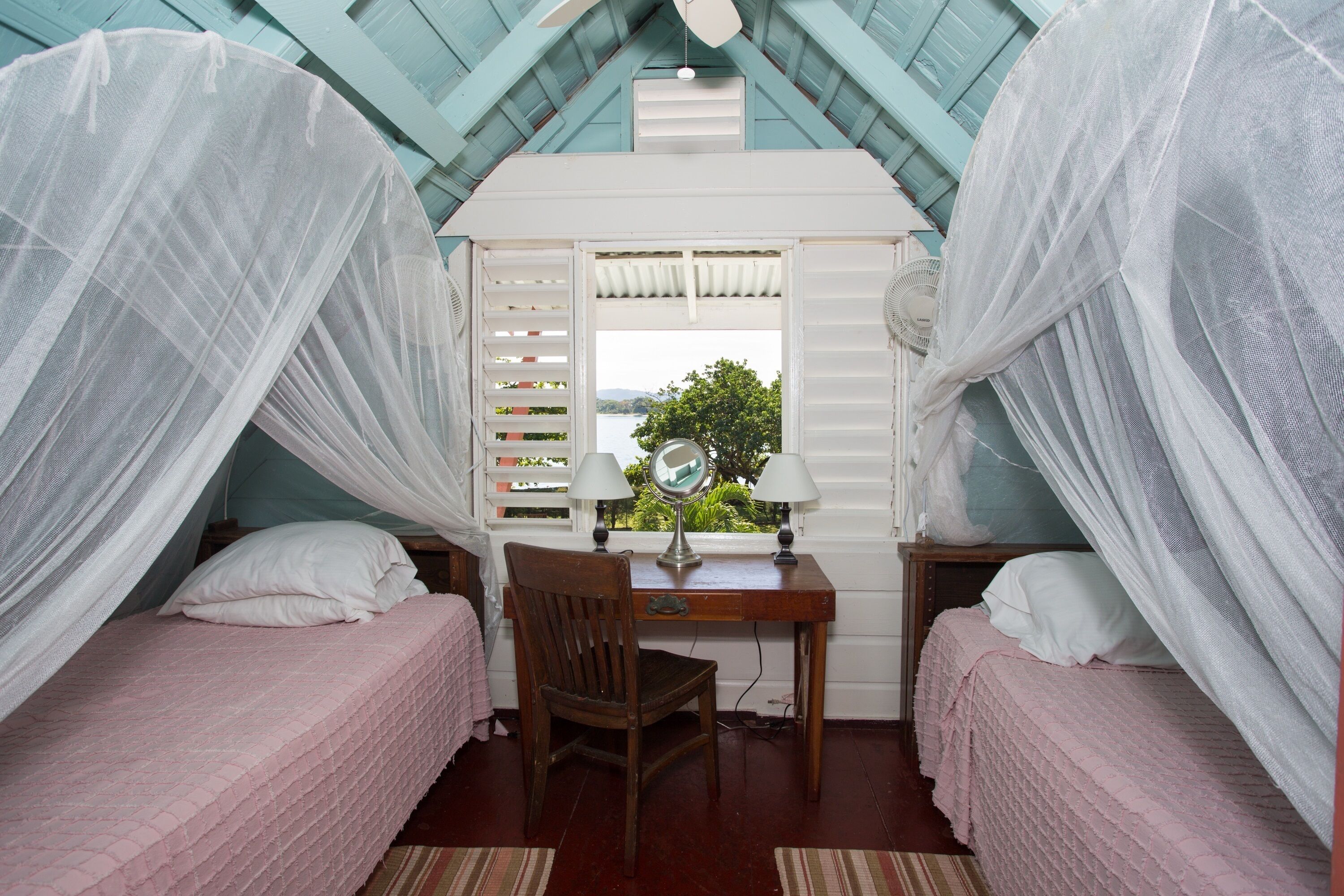 A bedroom with two beds, a desk with a mirror, and a window with a view of greenery.