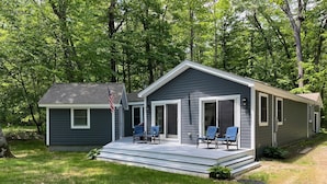 A small, single-story cabin with a porch and two chairs, surrounded by trees. - 3 bedroom, 2 Baths, 8 Sleeps, detached bunk house, open boat slip (Ossipee, NH 03814)