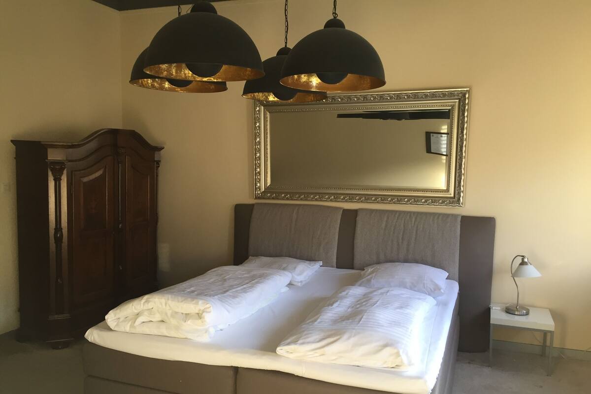 Un dormitorio con una cama grande, dos lámparas colgantes negras, un armario de madera, una mesita de noche con lámpara y un espejo sobre la cama.