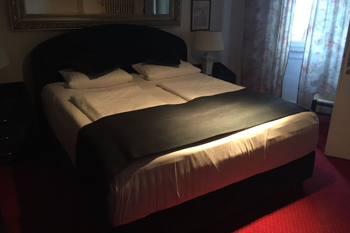 Una habitación de hotel con una cama grande, una mesita de noche, una lámpara y una ventana con cortinas.