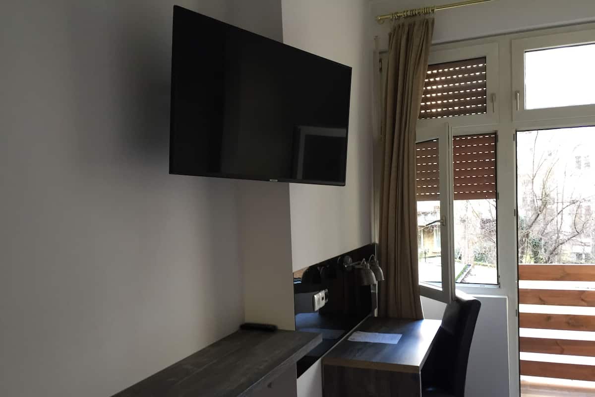 Habitación con televisión de pantalla plana en la pared, un escritorio de madera, una silla y una ventana con persianas.