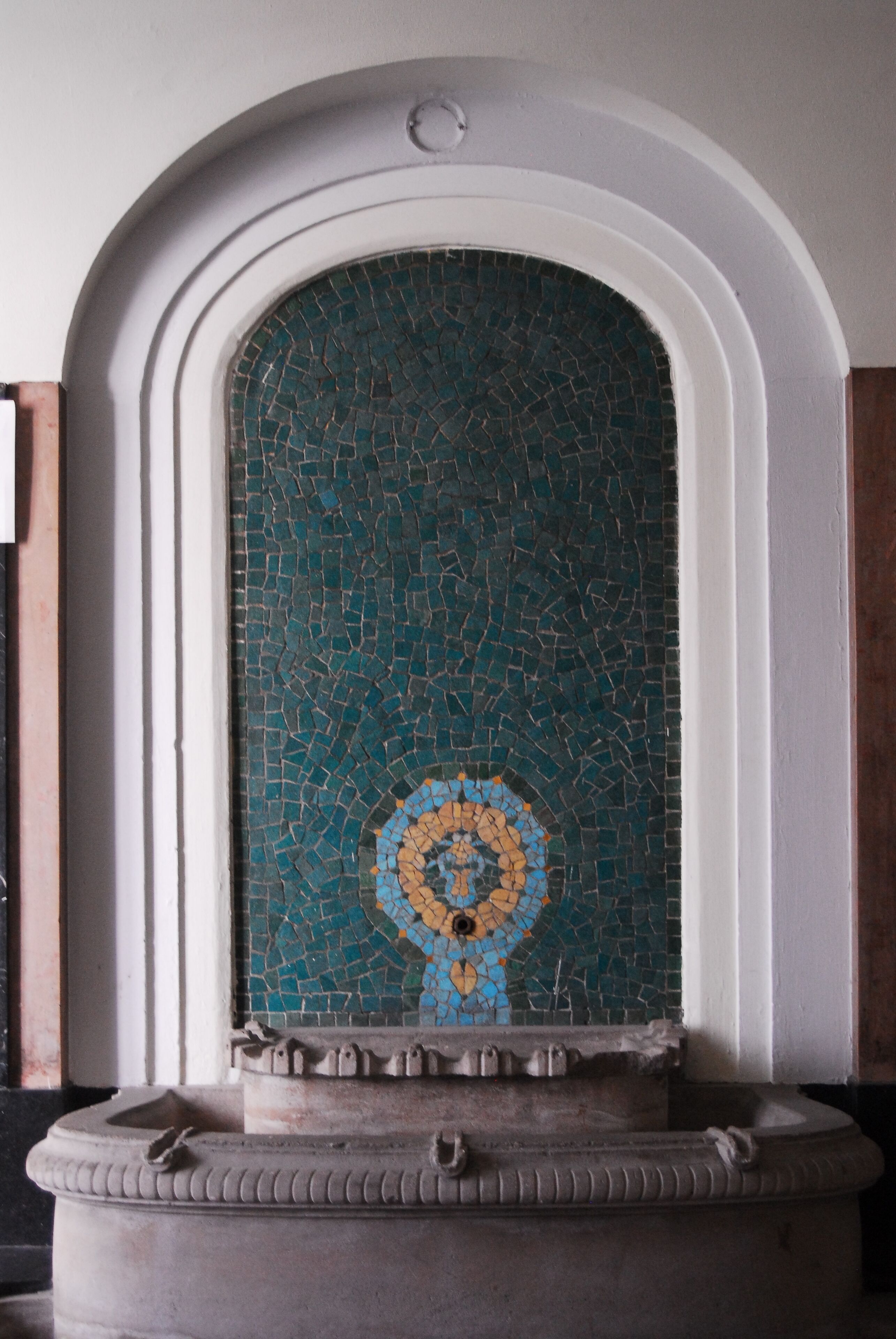A mosaic wall with a circular design, a stone base, and a white arch above.