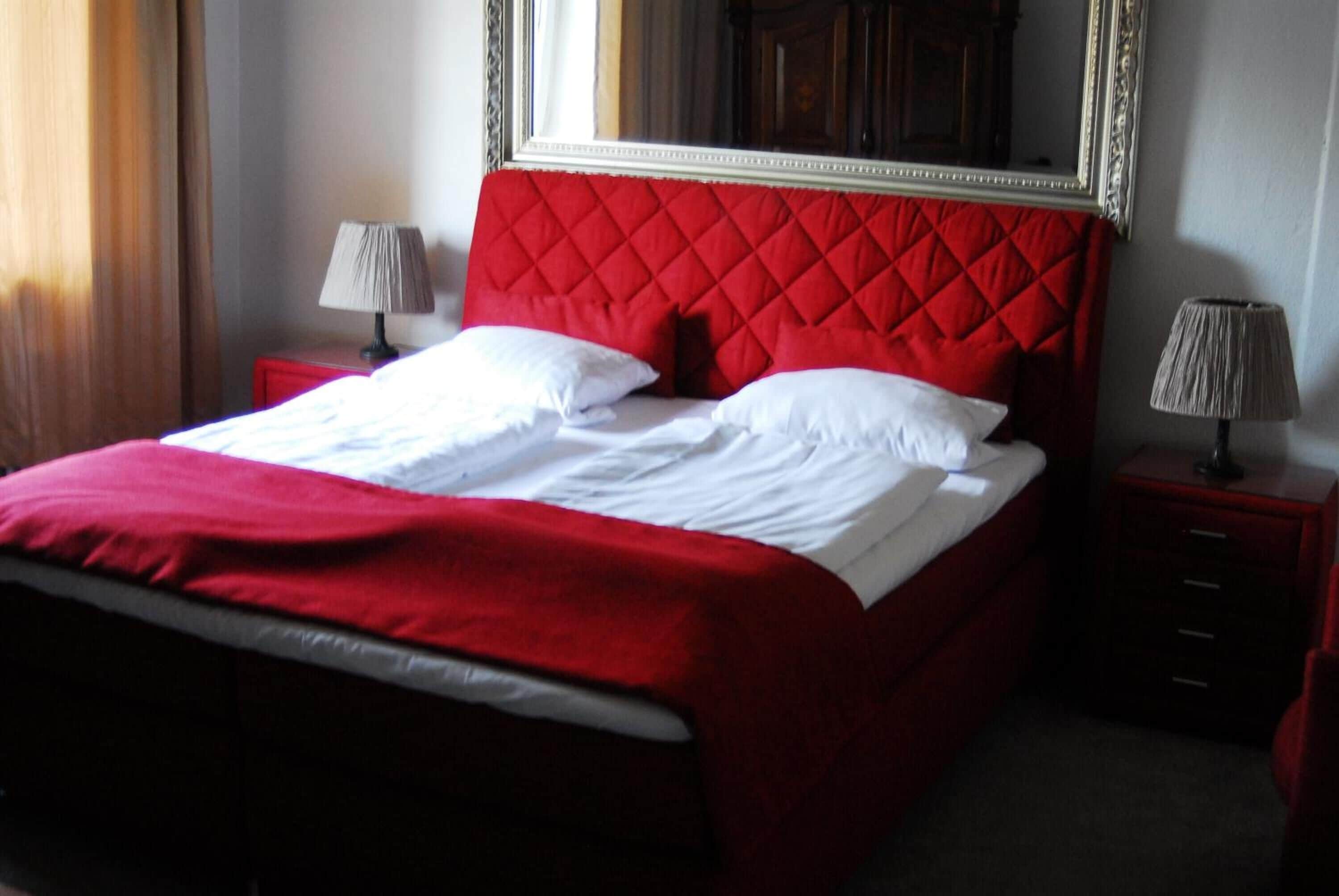 A neatly made bed with a red headboard and white bedding, two bedside tables with lamps, and a large mirror above the bed.
