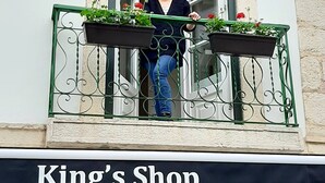 Una persona in piedi su un balcone con due fioriere, con vista su un edificio con un cartello che riporta la scritta 'King's Shop'.