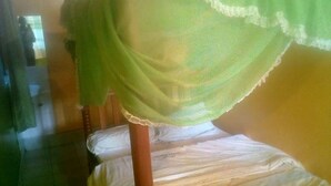 A single bed with a wooden frame and a white mattress. A green, sheer canopy is draped over the bed. The room has a tiled floor and a plain wall in the background. - Tripple Arrow Guest House (Nairobi)
