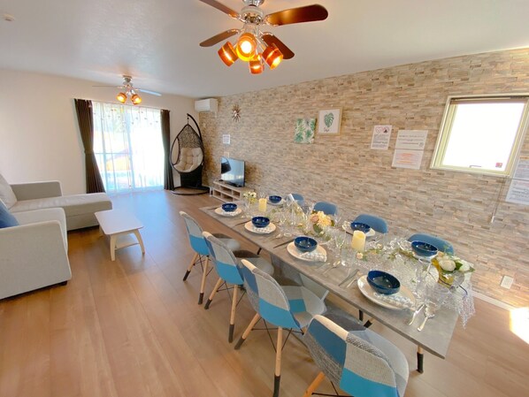 Un coin repas avec une longue table dressée pour six personnes, entourée de chaises bleues. Un téléviseur est posé sur un meuble à gauche, et un ventilateur de plafond est installé au-dessus.