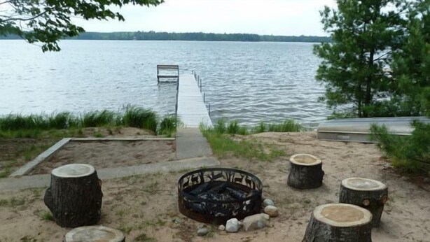 A lakeside setting with a fire pit surrounded by tree stumps, a dock extending into the water, and a sandy area with a concrete path.