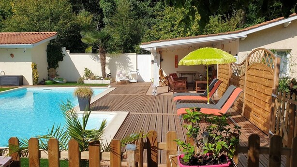 Un patio trasero con piscina, terraza de madera, sillas de descanso y zona de estar cubierta.