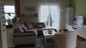 Una sala de estar con un sofá, una mesa de centro y un televisor. Hay una ventana con cortinas, una planta y una estantería con adornos.