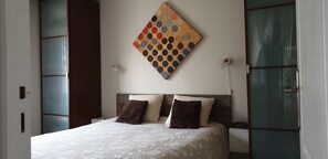 A neatly made bed with a patterned bedspread, a wall-mounted artwork, bedside lamps, and a nightstand with a clock. - House with swimming pool not overlooked- near Bordeaux - 3 bedrooms - 6 people (pian medoc)