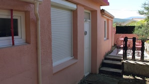 A pink building with a closed shuttered window, a door, and a set of steps leading to a patio area with outdoor furniture. - Independent studio in a quiet building with small patio (Vernet-les-Bains)
