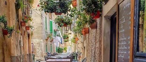 A narrow street with a table and chairs set up for outdoor dining, surrounded by buildings with hanging plants and a chalkboard menu.