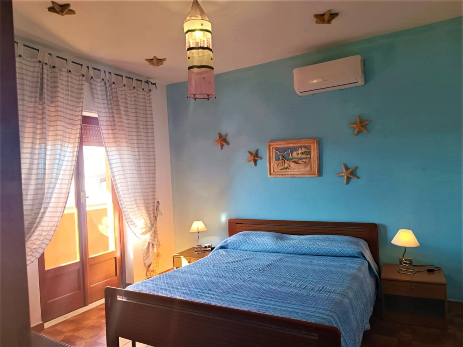 Un dormitorio con cama, dos mesitas de noche, lámpara colgante, aire acondicionado instalado en la pared y decoración con motivos de estrellas de mar.
