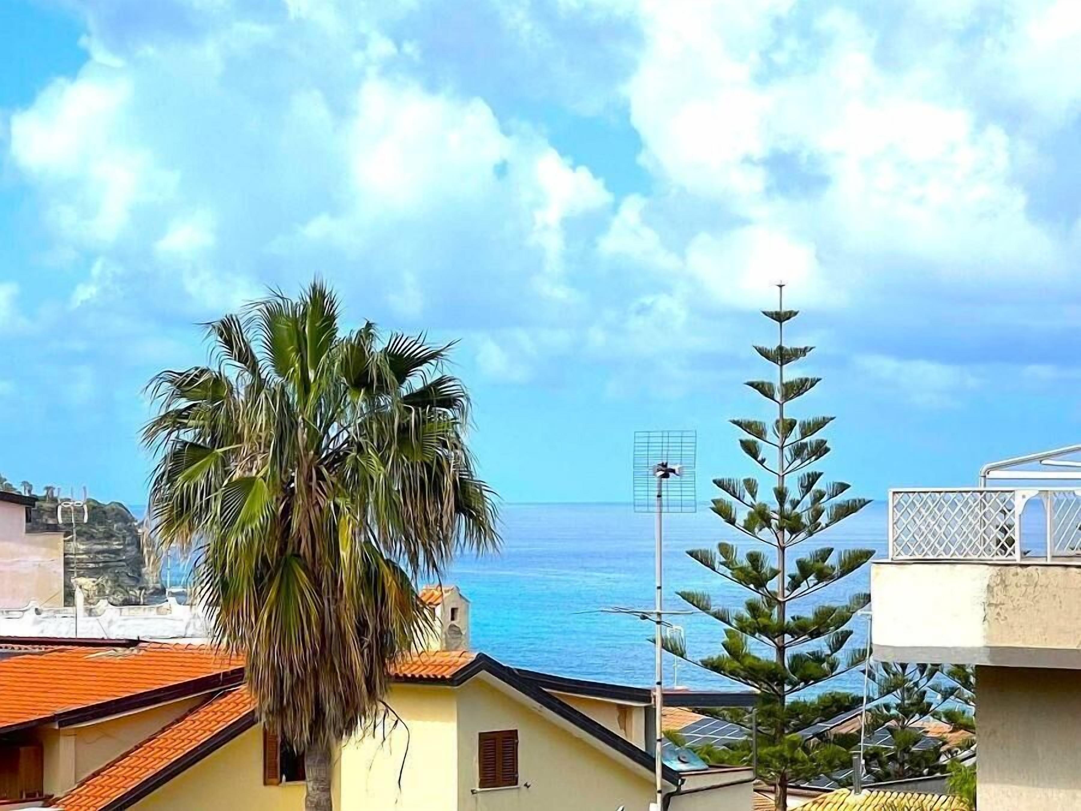 A coastal building with a view of the sea, a palm tree, and a pine tree.