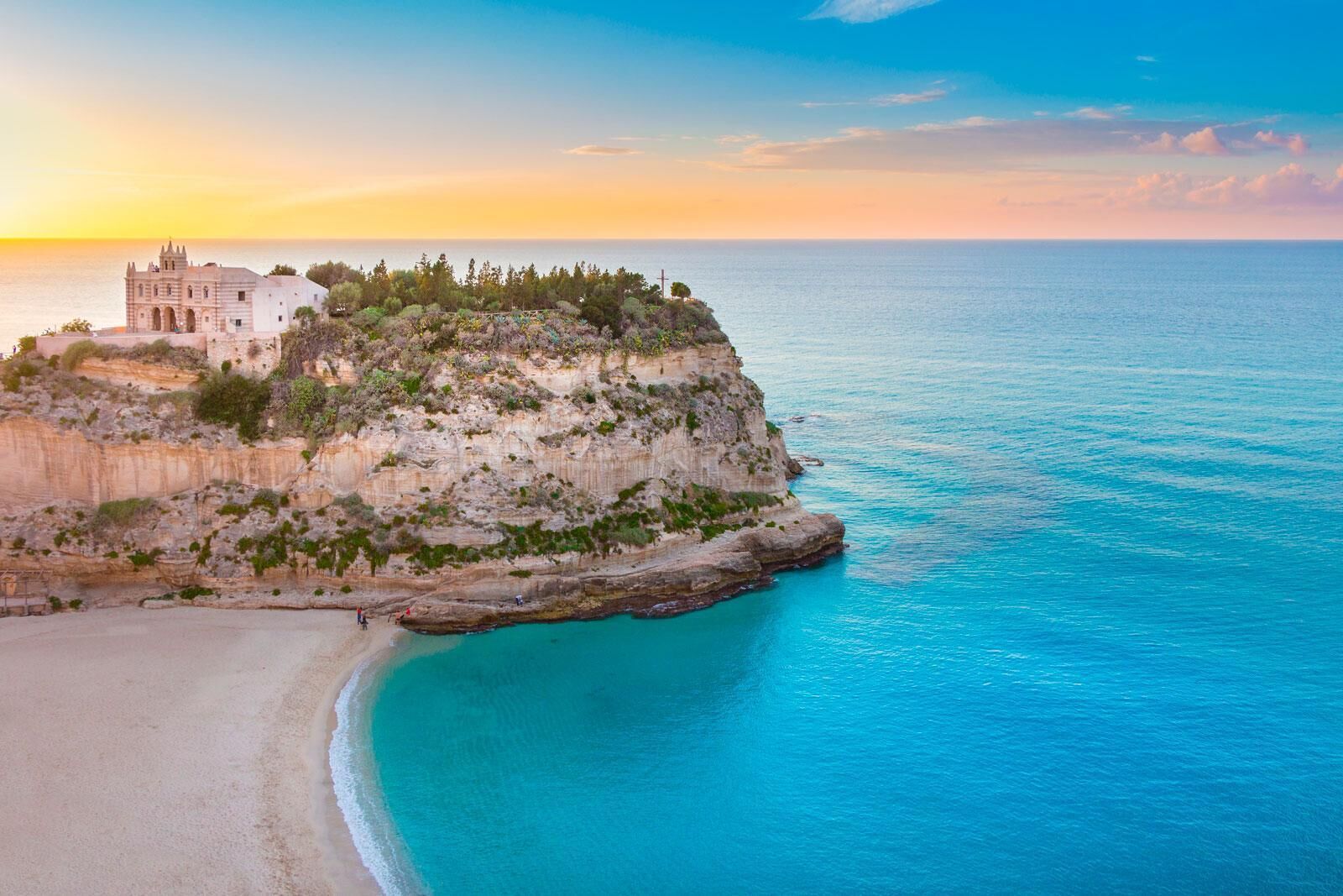 Edifici a la costa, amb una platja davant i un penya-segat amb vegetació.