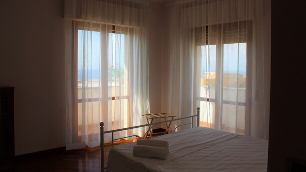 A bedroom with a bed, a chair, and large windows with sheer curtains. - Orchidea (Tropea)