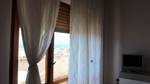 A room with a window, curtains, a television on a cabinet, and a view of a beach and buildings. - Orchidea (Tropea)
