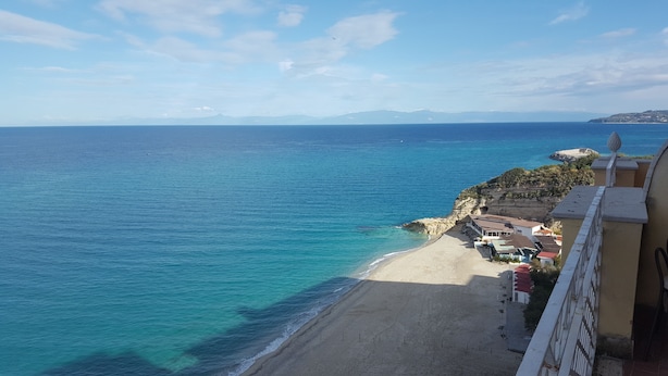 A coastal view with a beach, a building with a balcony, and a distant mountain range. - Sabbia (Tropea)