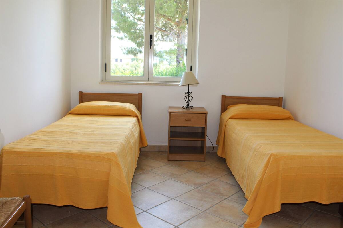 Quarto com duas camas, uma mesa de cabeceira e janela com vista para árvores.