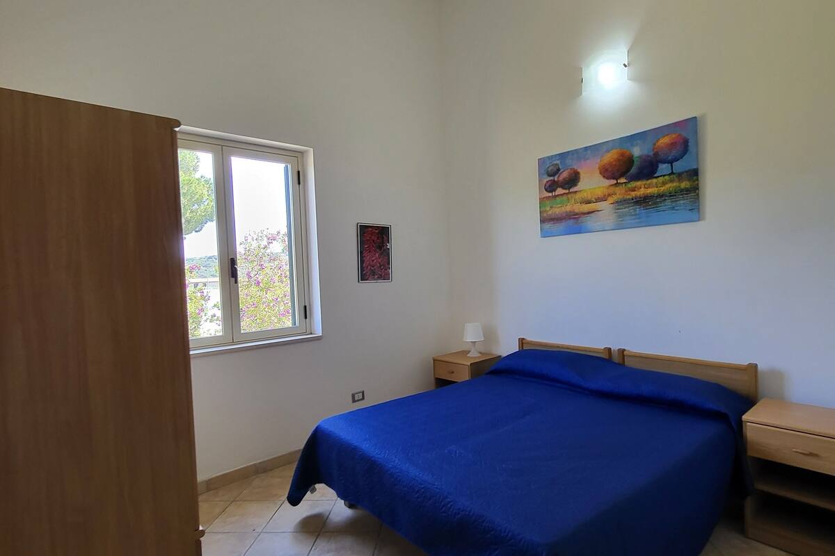 Um quarto com uma cama, uma mesa de cabeceira, um guarda-roupa de madeira, uma janela e um quadro na parede.