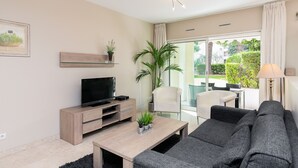 A modern living room with a flat-screen TV, a coffee table with plants, a grey sofa, and a large window with curtains.