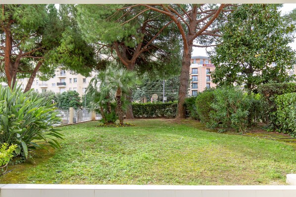 A courtyard with a grassy lawn, several trees, and a building with balconies in the background. - 2 Bed - Sleeps 5 - Pool Access - Central Cannes (Cannes)