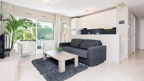 A modern living room with a grey sofa, a wooden coffee table, and a black rug. There is a kitchen area with white cabinets and a black countertop.