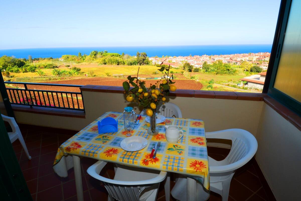 Un balcon avec une table dressée pour deux personnes, un vase rempli de citrons et une vue sur la mer et la ville.
