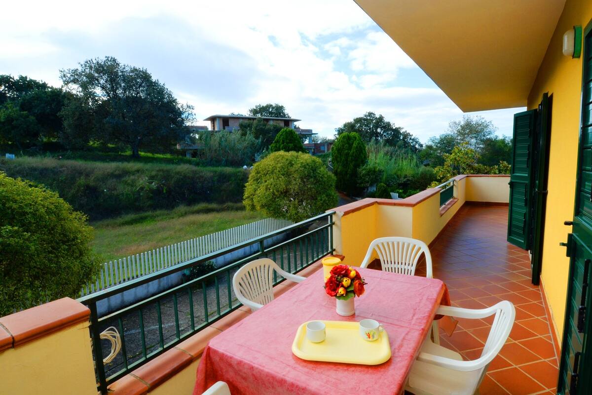 Un balcon avec une table dressée pour deux personnes, un vase de fleurs et une vue sur un paysage verdoyant.