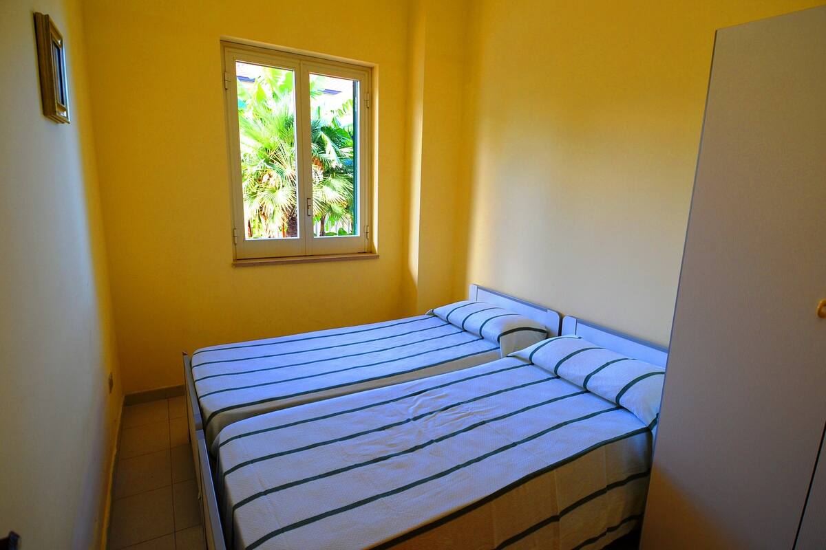 Une petite chambre avec un lit simple, une fenêtre, des murs unis et une porte.