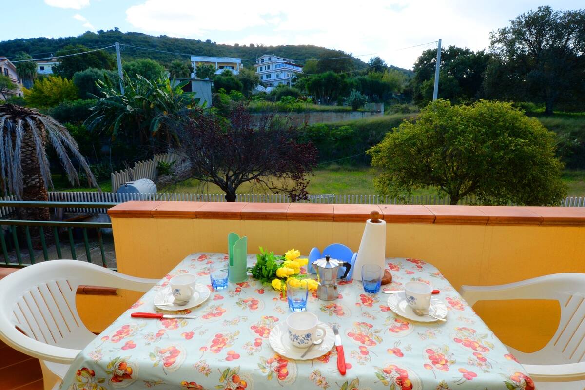Une table de balcon décorée avec une nappe à motifs floraux, des assiettes, des tasses et un arrangement floral.