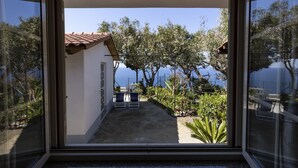 Vista a través de un ventanal corredero de una casa con patio, una pequeña mesa y sillas, con vista a un jardín y una masa de agua.