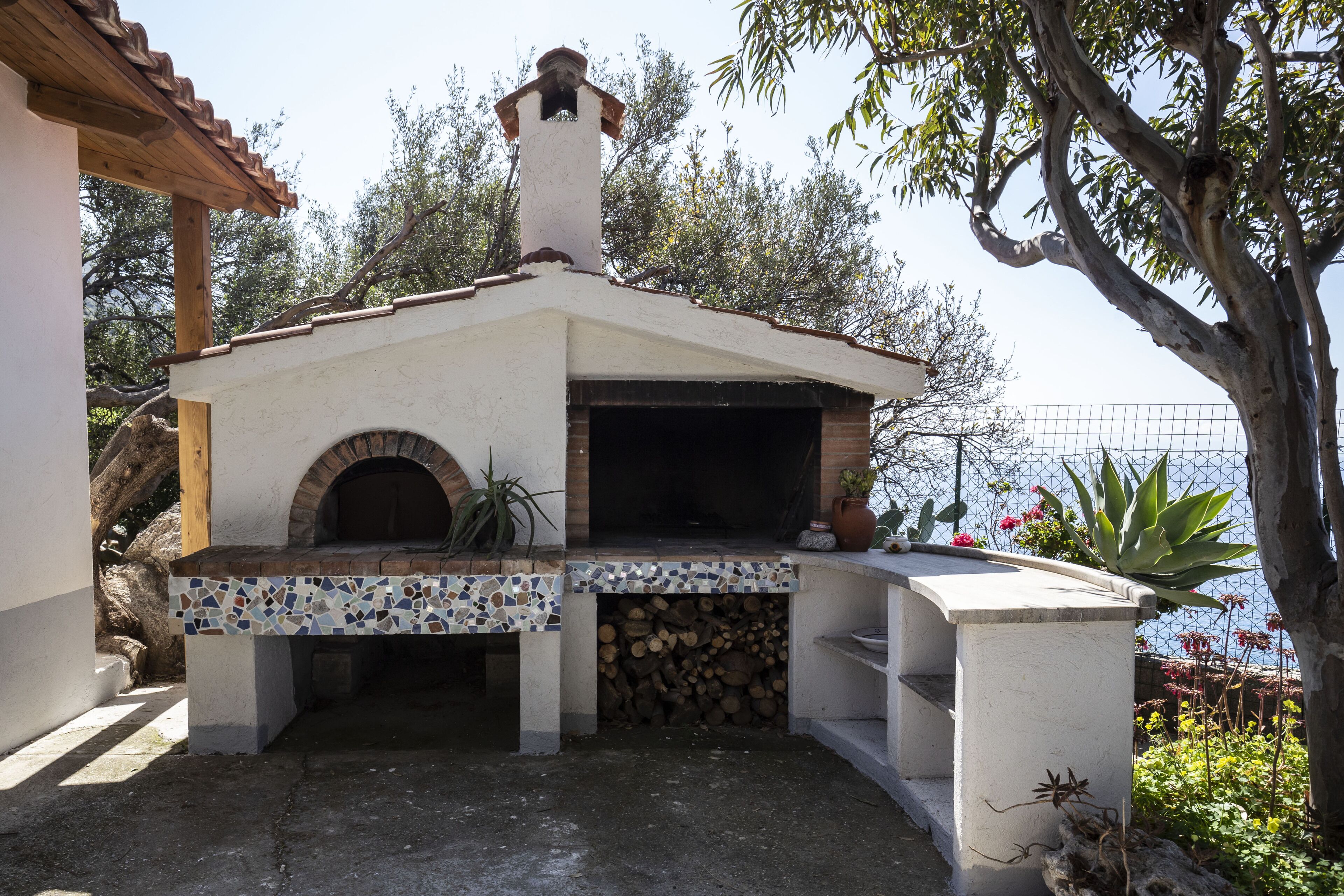 An outdoor brick oven with a tiled base, surrounded by plants and trees.