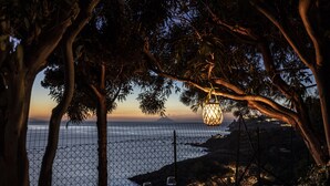 Mesa con dos copas de vino y una vela, con vista a una zona costera con cerca y árboles.
