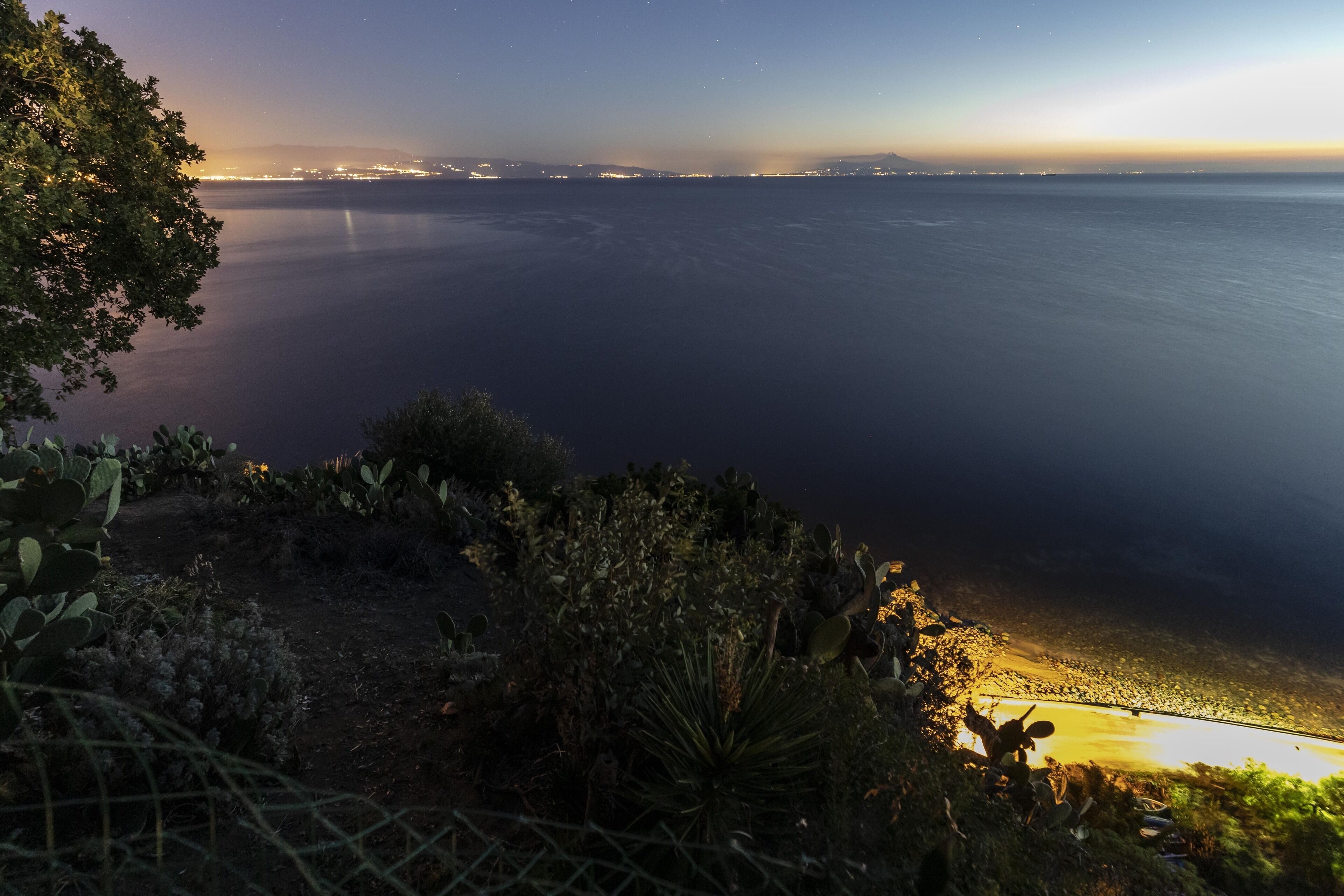 A coastal landscape with a view of the sea and distant city lights.