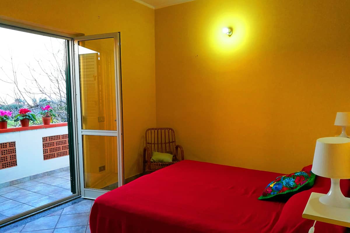 A room with a red bed, a chair, a small table with a lamp, and a window with potted plants.