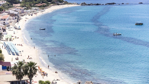 Una playa costera con aguas azules cristalinas, orilla de arena y algunas embarcaciones.