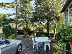 Une terrasse en pierre avec une table et des chaises blanches, entourée d’une végétation luxuriante et d’arbres.