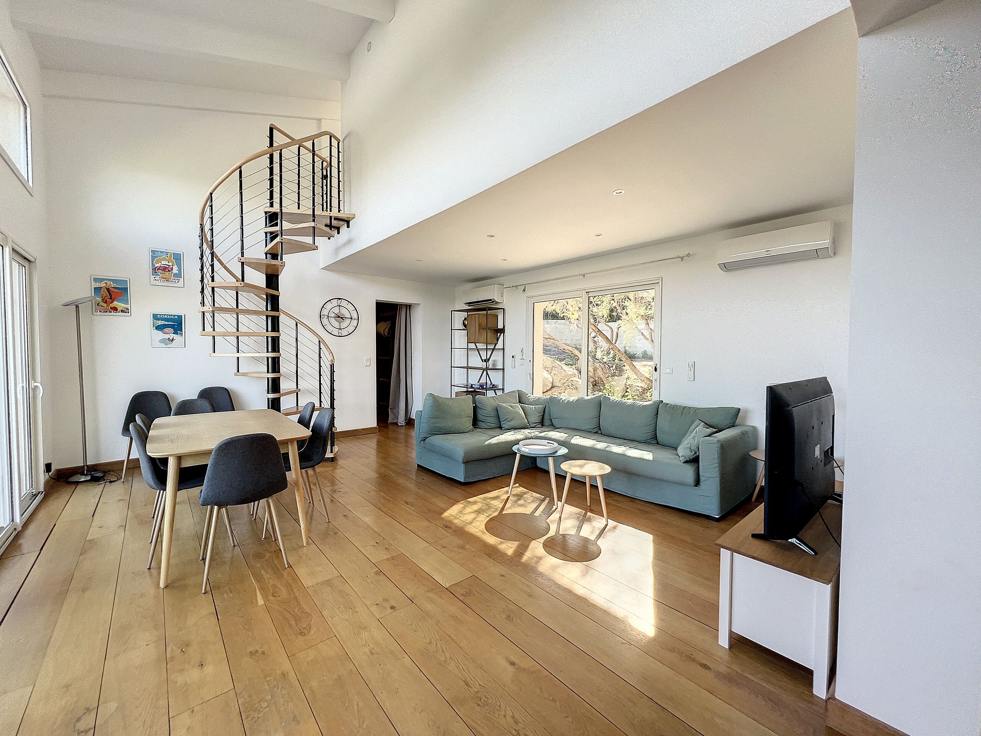A spacious living area with a wooden floor, a spiral staircase, a sofa, a dining table with chairs, and a television.