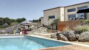 En swimmingpool med solstolar och en möblerad, överbyggd sittplats, omgiven av stora stenar och en byggnad med stora fönster.