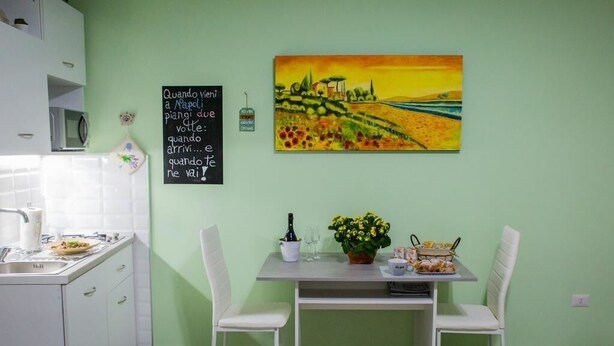 Una cocina con armarios blancos, una mesa de comedor con dos sillas blancas, un cuadro en la pared y una pizarra con texto.