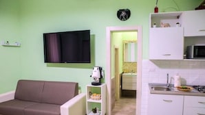 Una sala de estar con un sofá, un televisor, un estante pequeño con una lámpara y adornos, y una zona de cocina con armarios y un microondas.