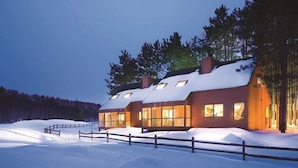 A modern, two-story cabin with large windows, surrounded by snow-covered ground and trees.
