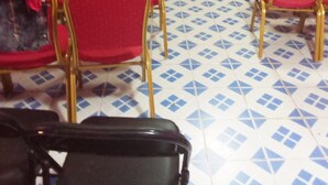 A room with rows of red chairs with gold frames, a black folding chair, and a blue table in the center. The floor has a blue and white geometric pattern.