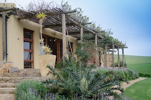 Uma casa com fachada de pedra, pérgula de madeira e um jardim bem cuidado, com diversas plantas e um caminho de pedras.