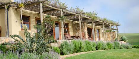 Une maison à deux étages dotée d’une pergola au-dessus de la terrasse, entourée d’un jardin bien entretenu avec divers arbustes et une pelouse.